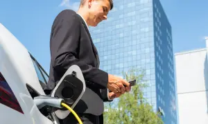 Uomo in giacca e cravatta controlla il telefono mentre la sua auto elettrica si ricarica, pianificare viaggio auto elettrica è facile con la tecnologia.