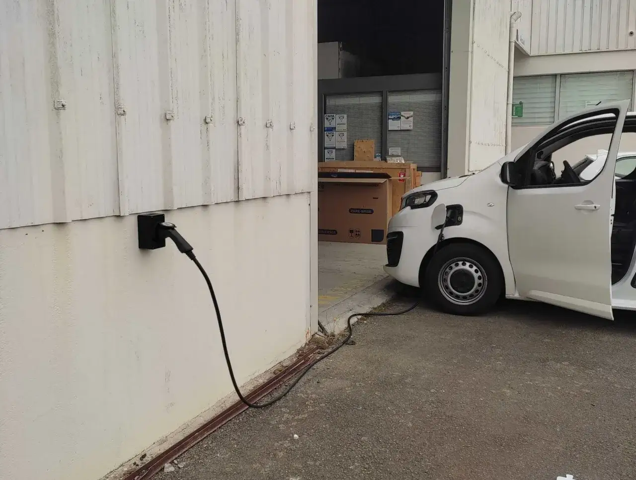 Un'auto elettrica bianca è in carica vicino a un edificio industriale. Un cavo nero collega la presa a muro al veicolo, pronto per un pieno di energia, un vero bonus per la mobilità elettrica.