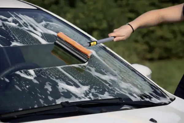 Mano che lava il parabrezza di un'auto con una spugna e schiuma, per imparare come pulire i vetri senza aloni.